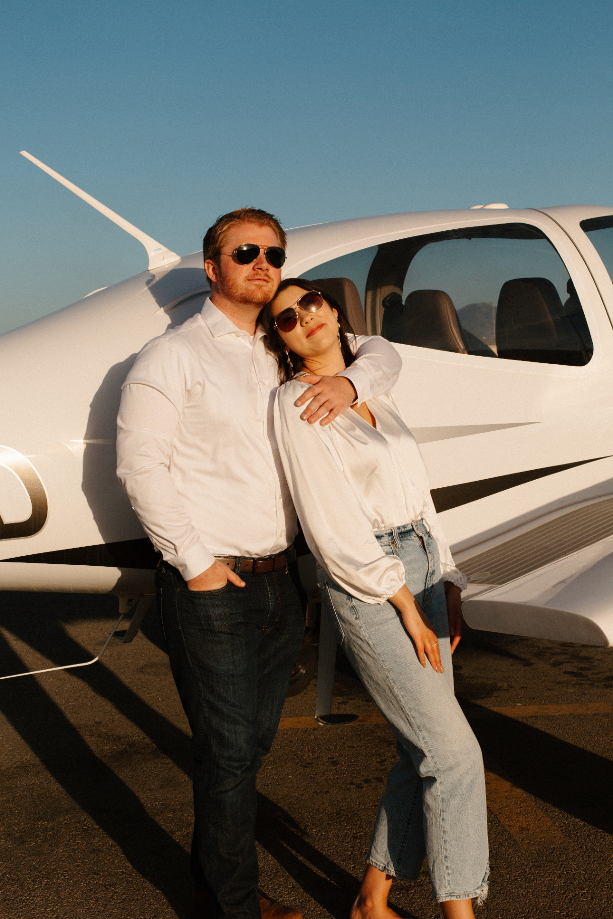 Couple in white shirts and sunglasses leaning against each other in front of a small white airplane on the tarmac at golden hour