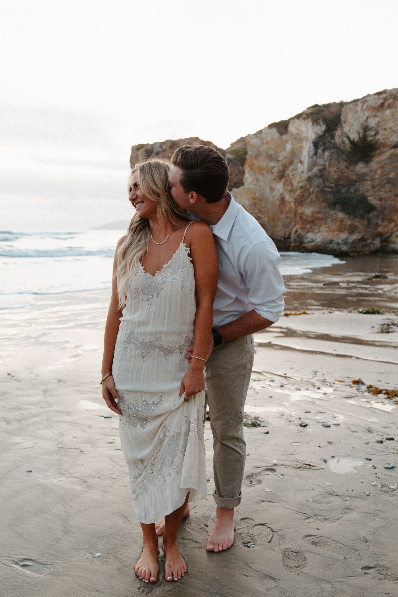 Groom in a white shirt kissing his smiling bride's neck as she holds her beaded ivory slip dress on a sandy beach with sandstone cliffs at golden hour