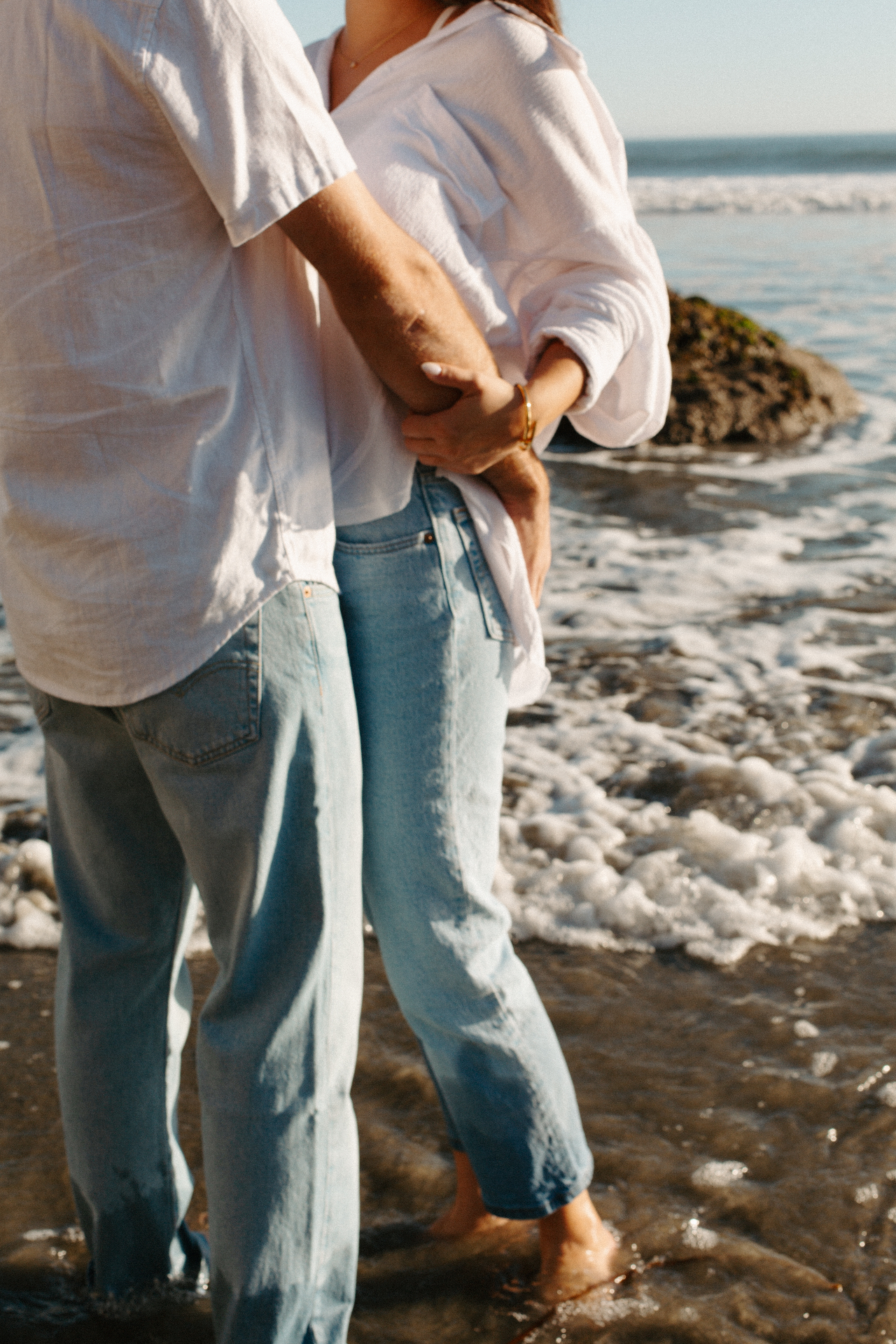 Cropped image of a couple in white shirts and light denim jeans embracing barefoot in the surf beside a mossy tide pool rock