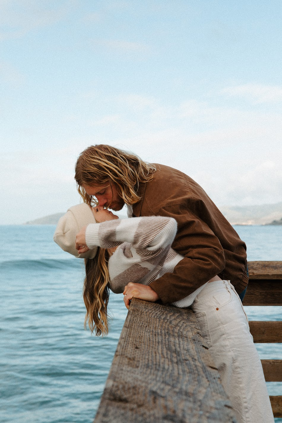 Couple kissing over a wooden pier railing above calm ocean water