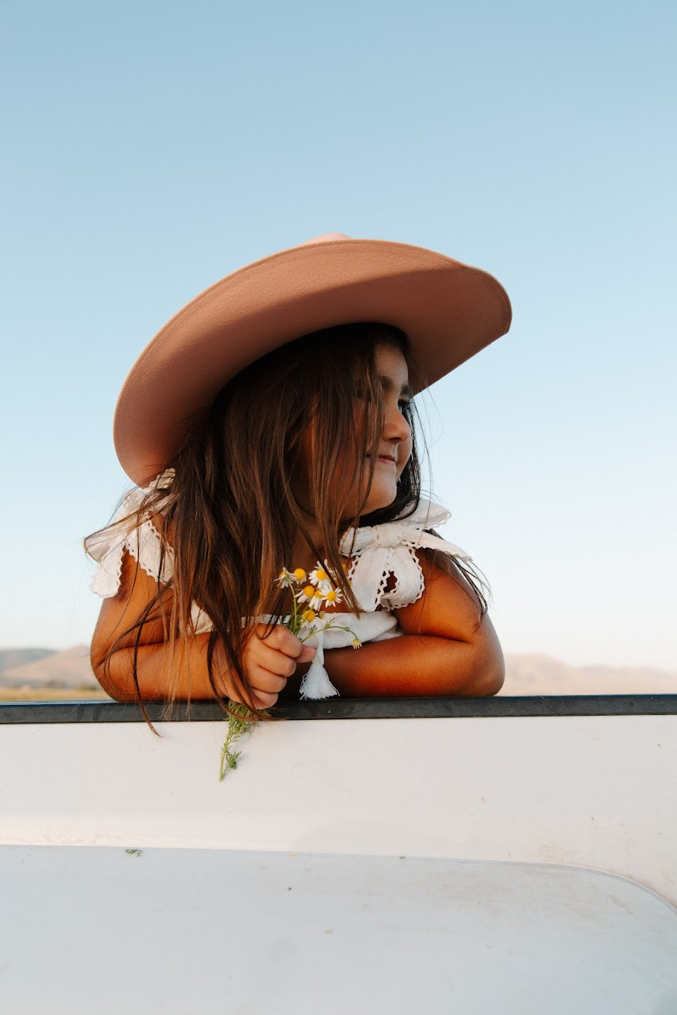 Little girl in a cowgirl hat holding wildflowers at golden hour