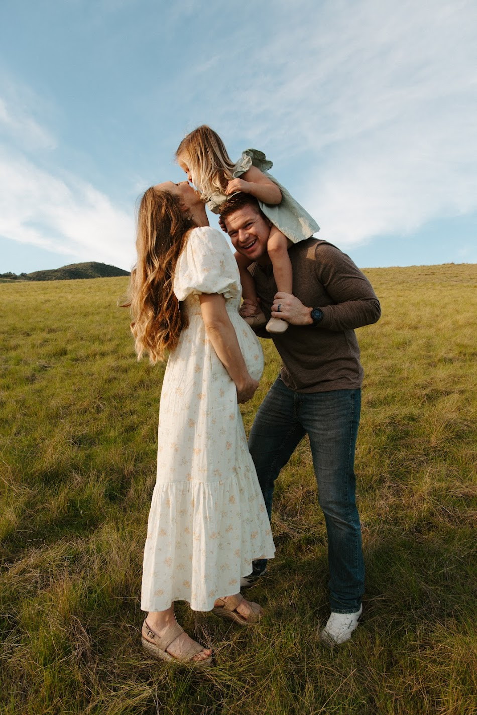 Mother kissing daughter on dad's shoulders in a grassy meadow