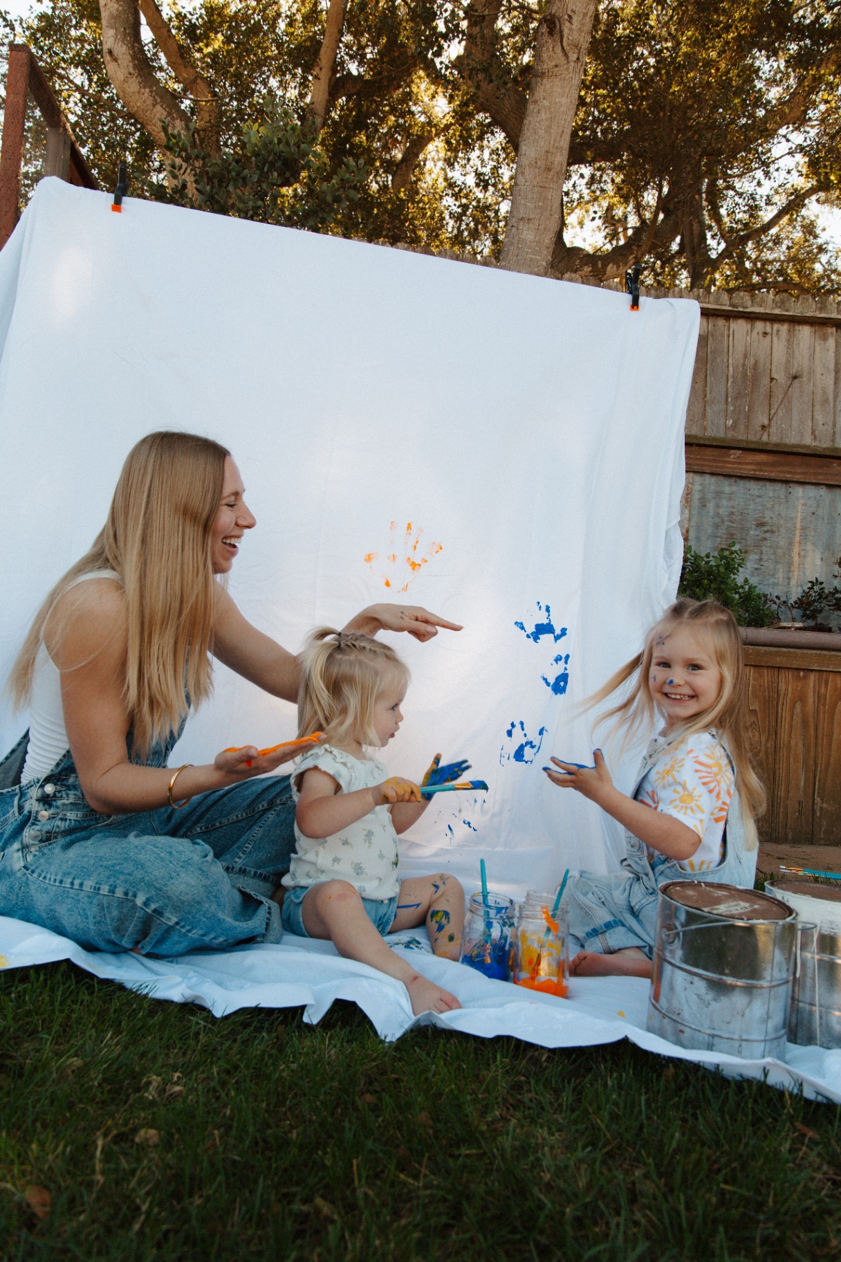 Mother and two daughters laughing while finger-painting handprints on a backyard canvas at golden hour