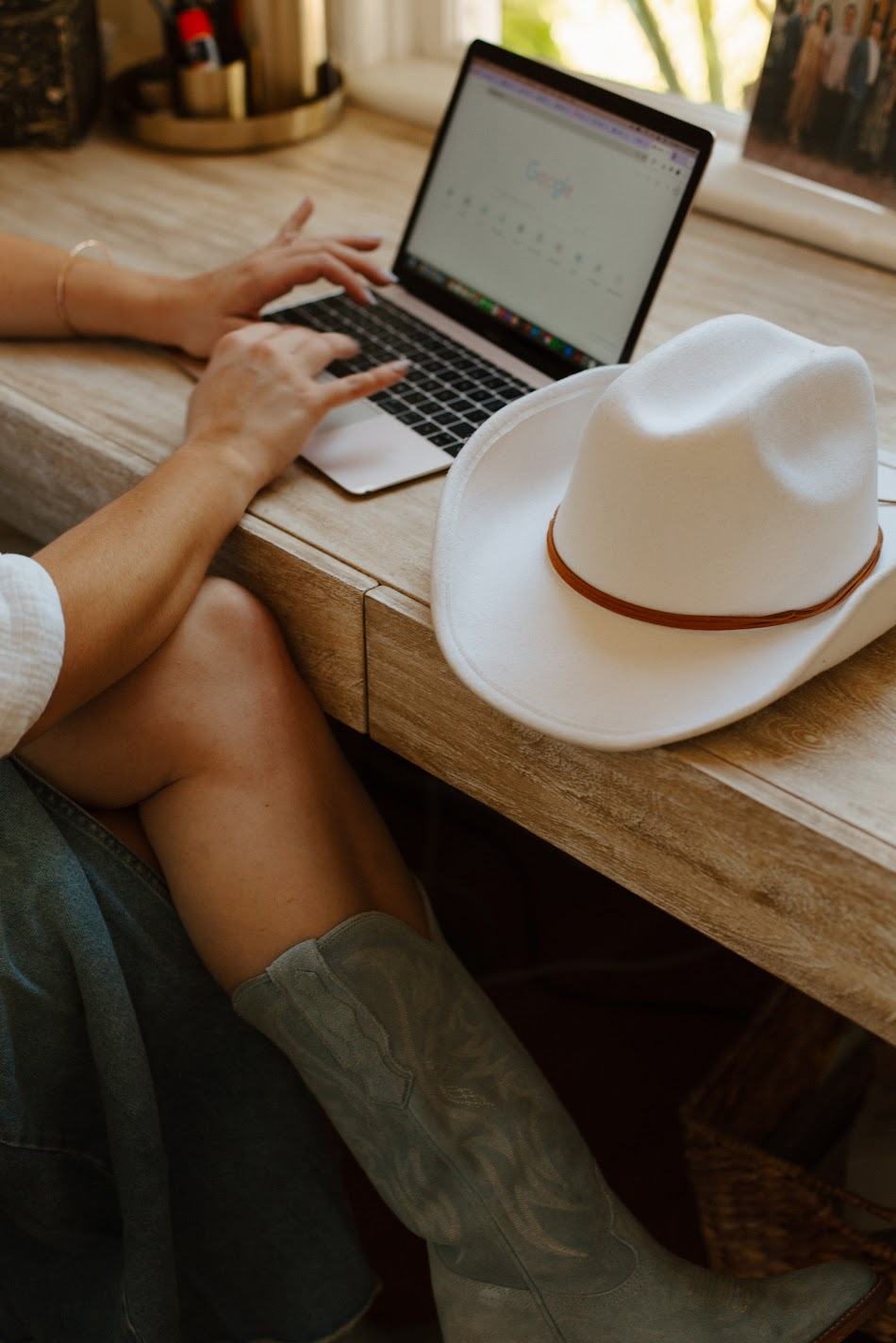 Woman working at a wooden desk with a laptop, white cowboy hat, and suede boots