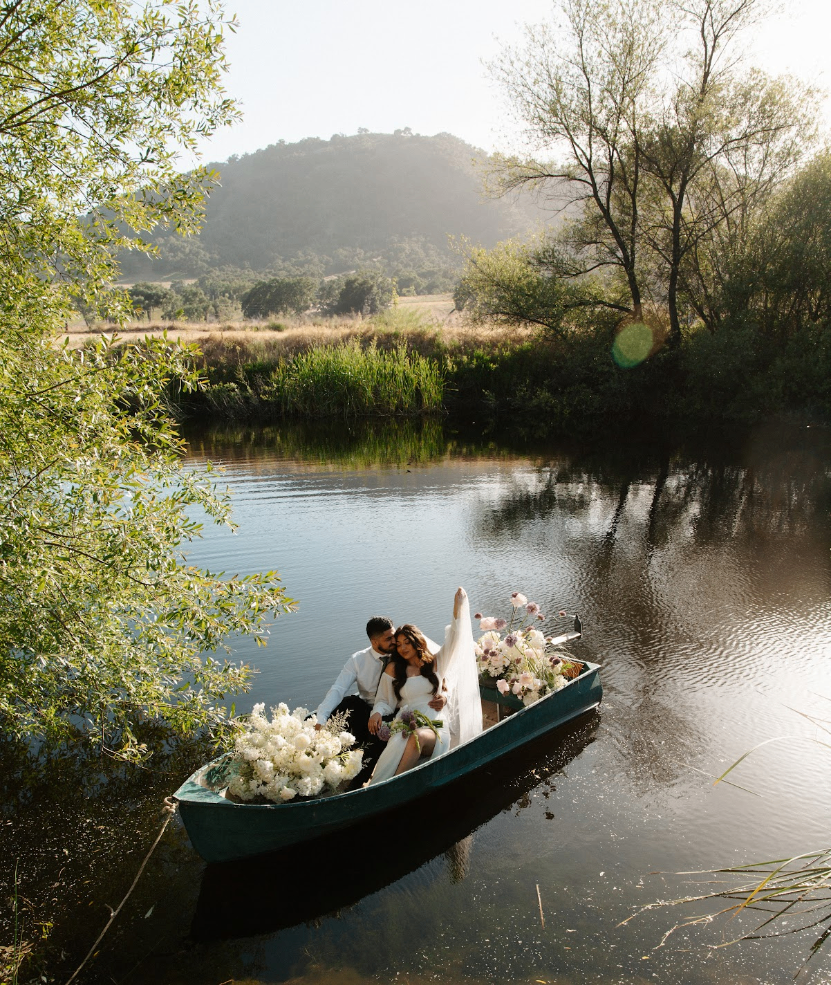 Bride and groom seated in a wooden rowboat with bouquets, on a still river at golden hour