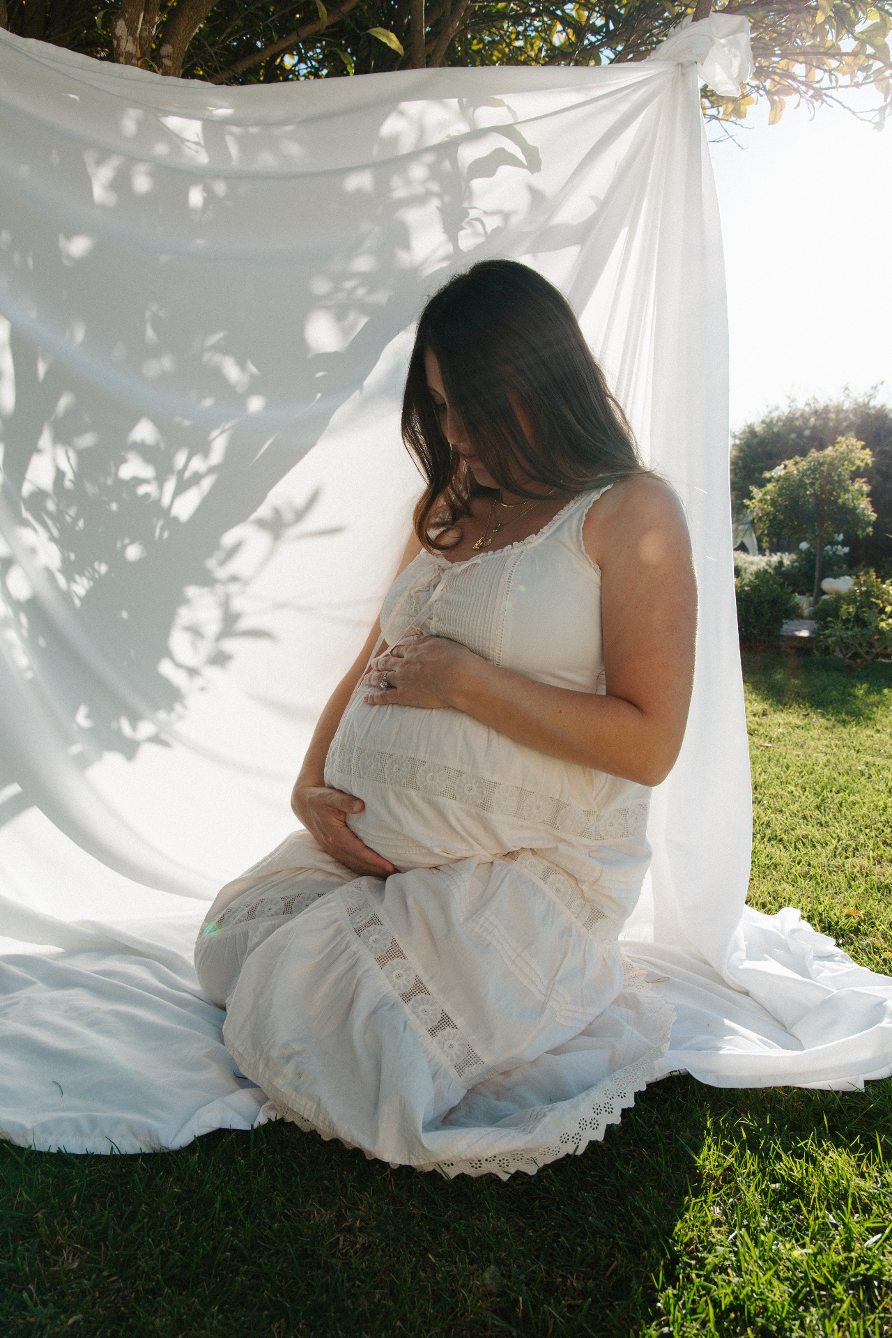Expecting mother in a lace white dress cradling her belly while seated on grass against a backlit sheer veil hung from a tree