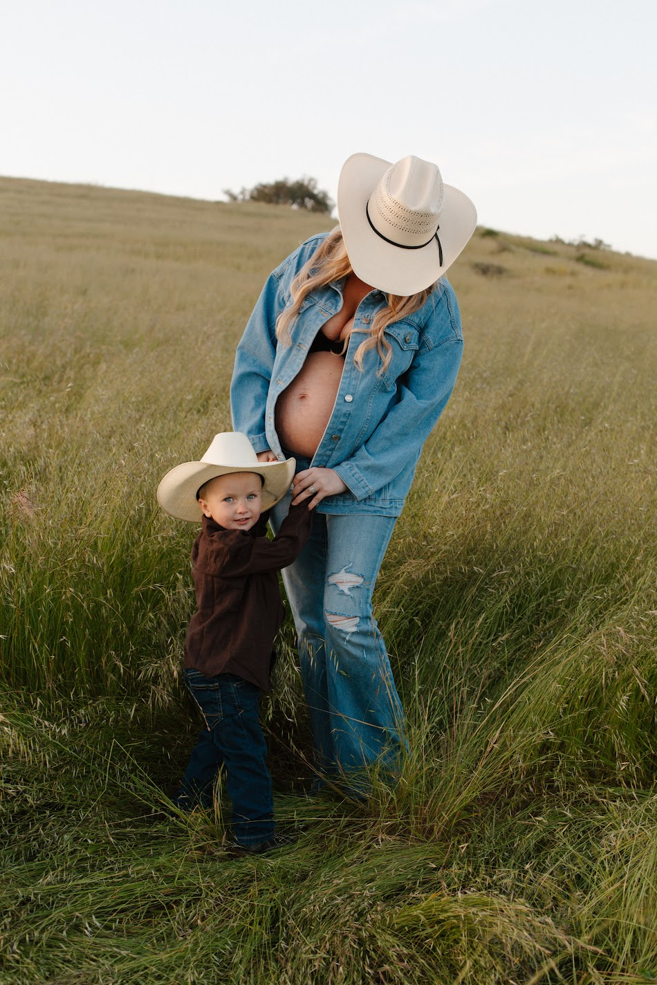 Expecting mother in a denim jacket and cowboy hat with her son in a grassy field