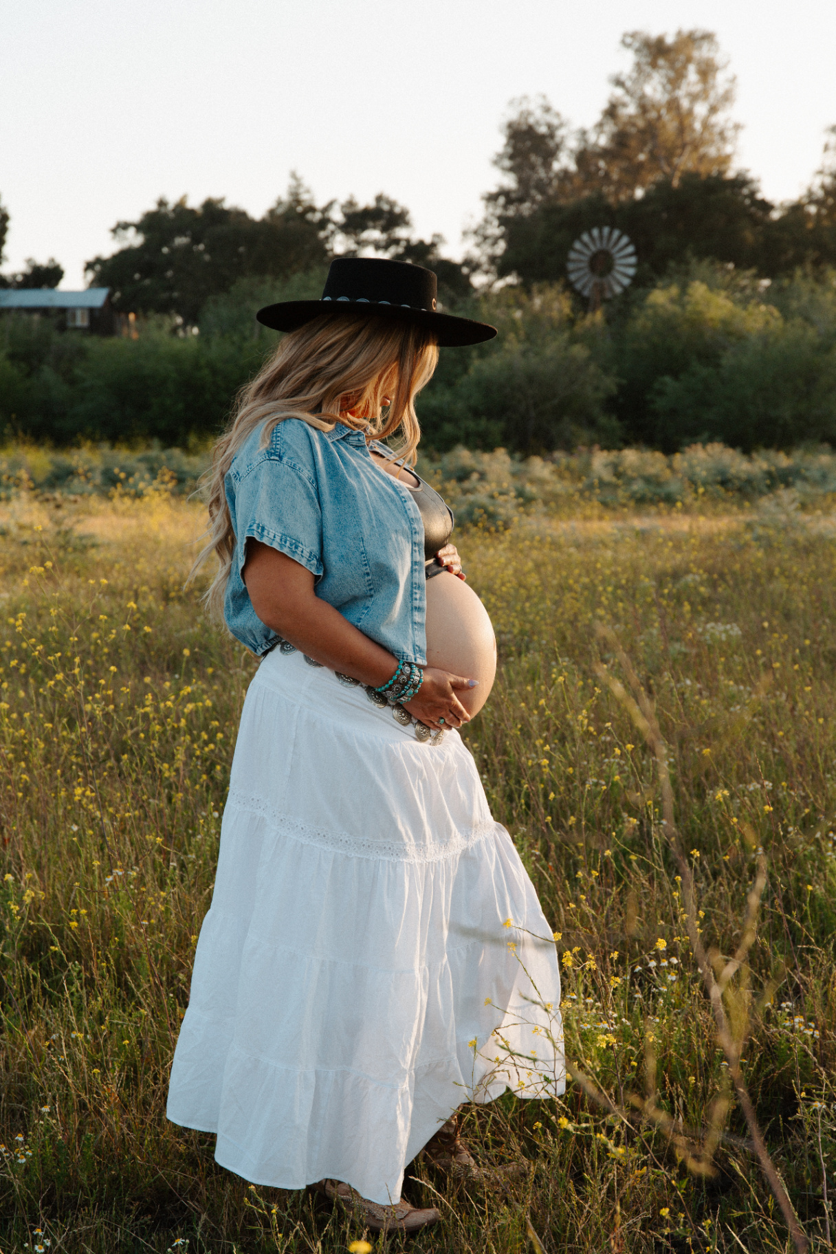 Expecting mother in a black wide-brim hat, denim shirt and flowing white skirt cradling her belly in a wildflower field with a windmill at golden hour