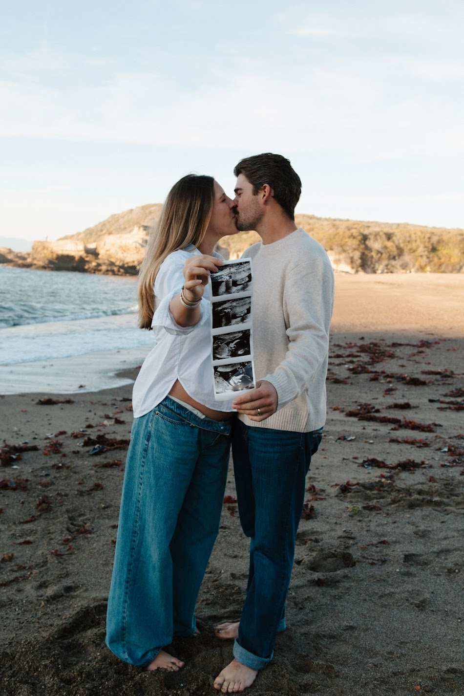 Expecting couple kissing on a sunset beach while holding up sonogram photos