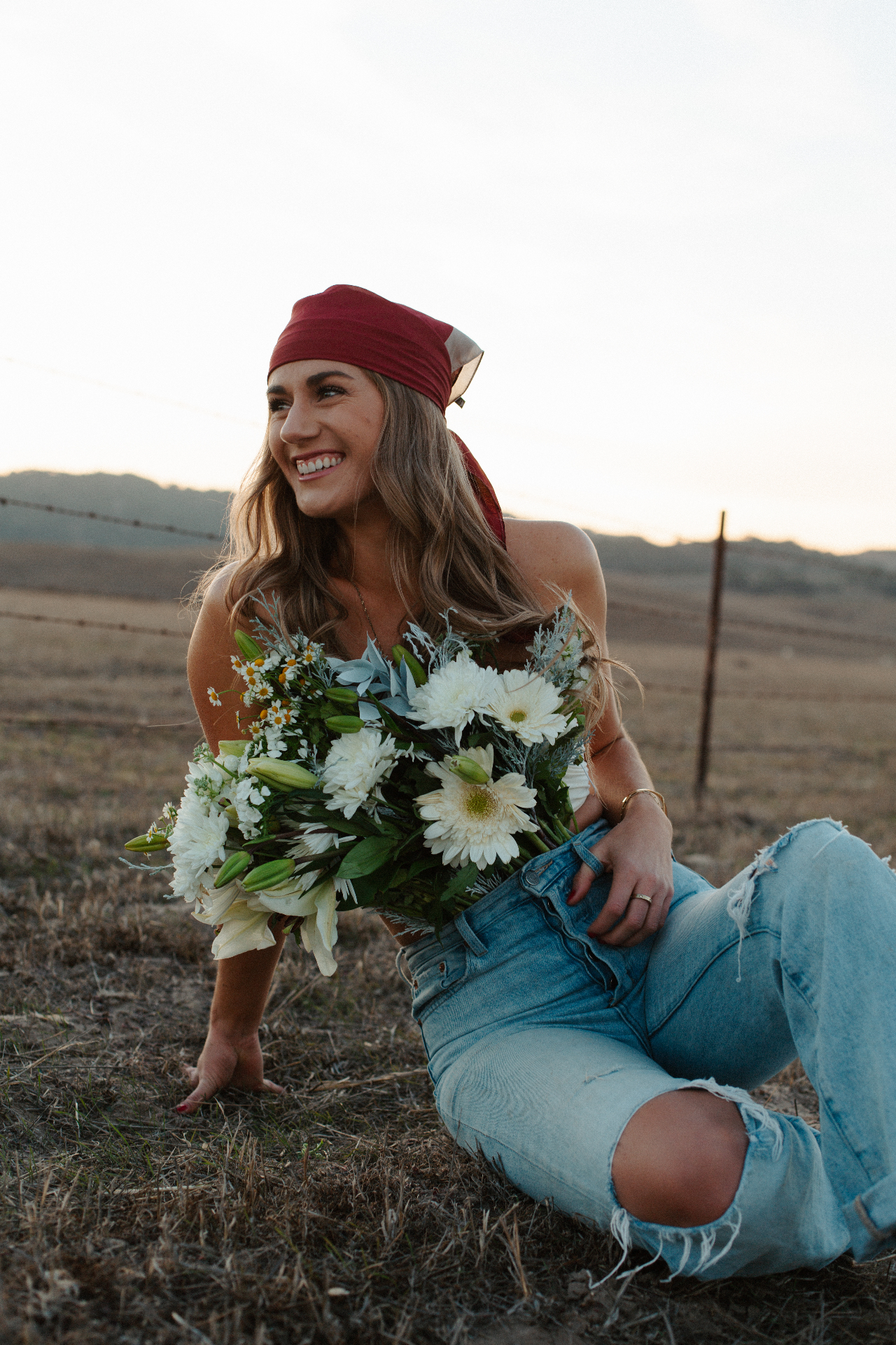 Young woman in a red bandana and ripped jeans laughing while seated in a sunlit field holding a white wildflower bouquet at golden hour
