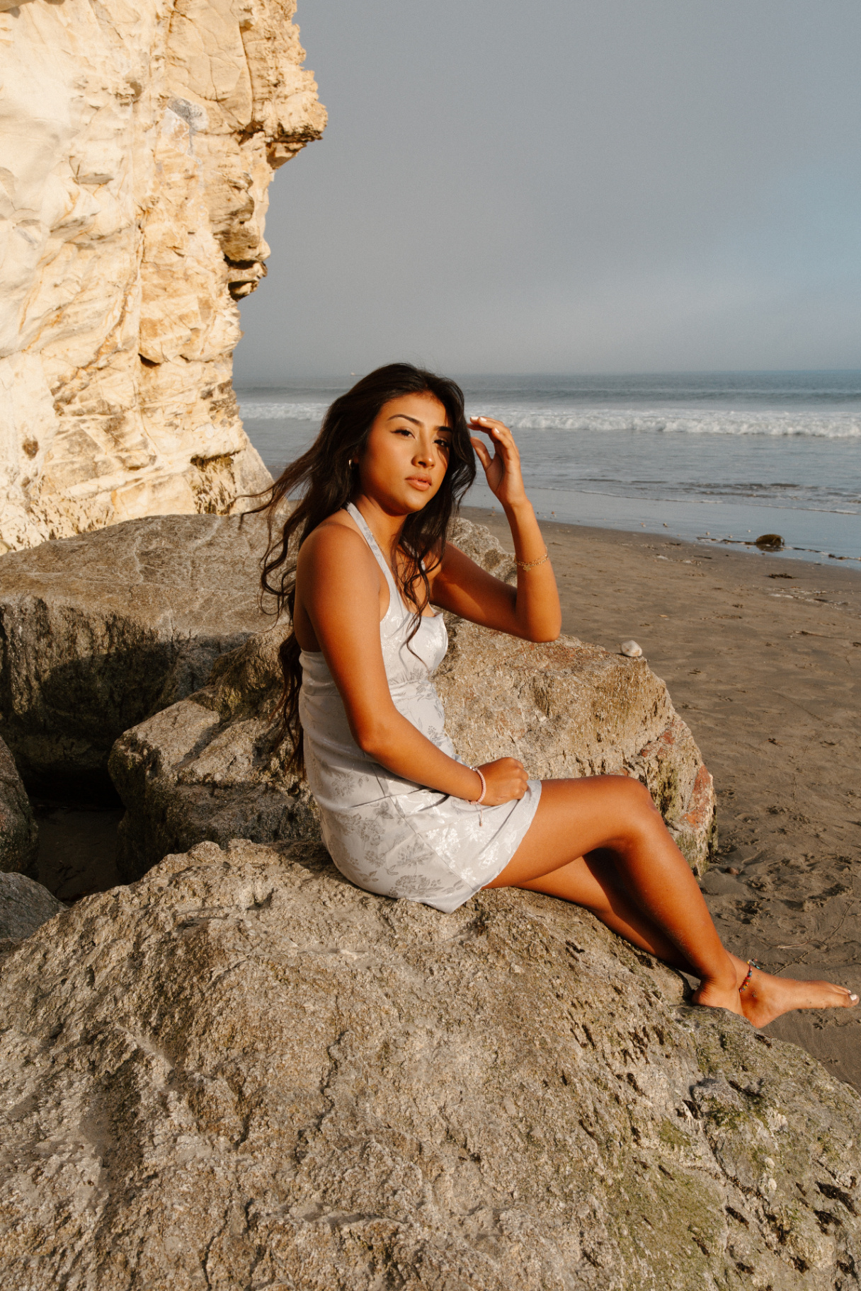Young woman in a silver floral mini dress seated on a large rock at the base of sandstone cliffs with the ocean behind at golden hour