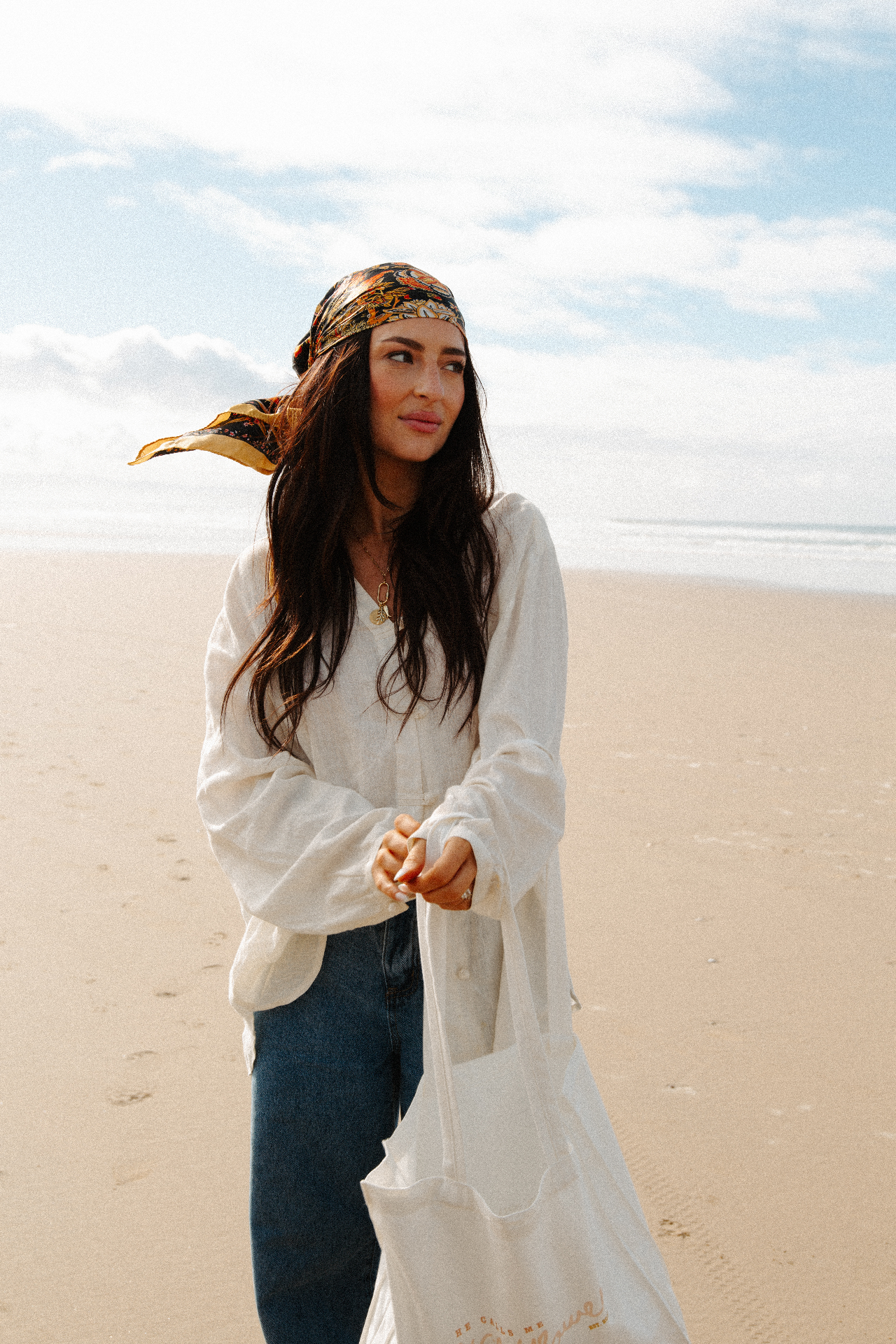 Woman in a printed silk headscarf and flowing white blouse holding a canvas tote on a wide sunlit beach