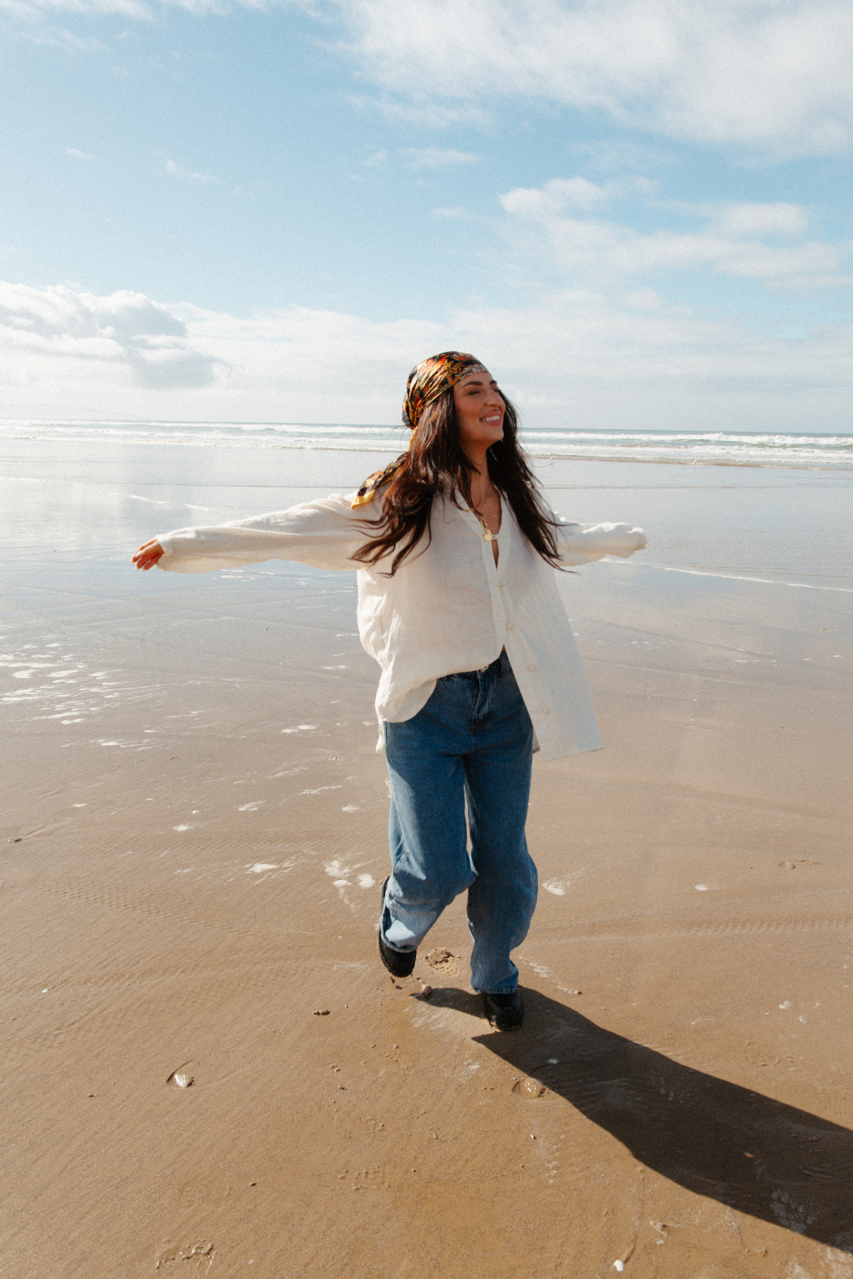 Woman in a printed headscarf, cream cardigan and blue jeans twirling barefoot on a wide sunlit beach