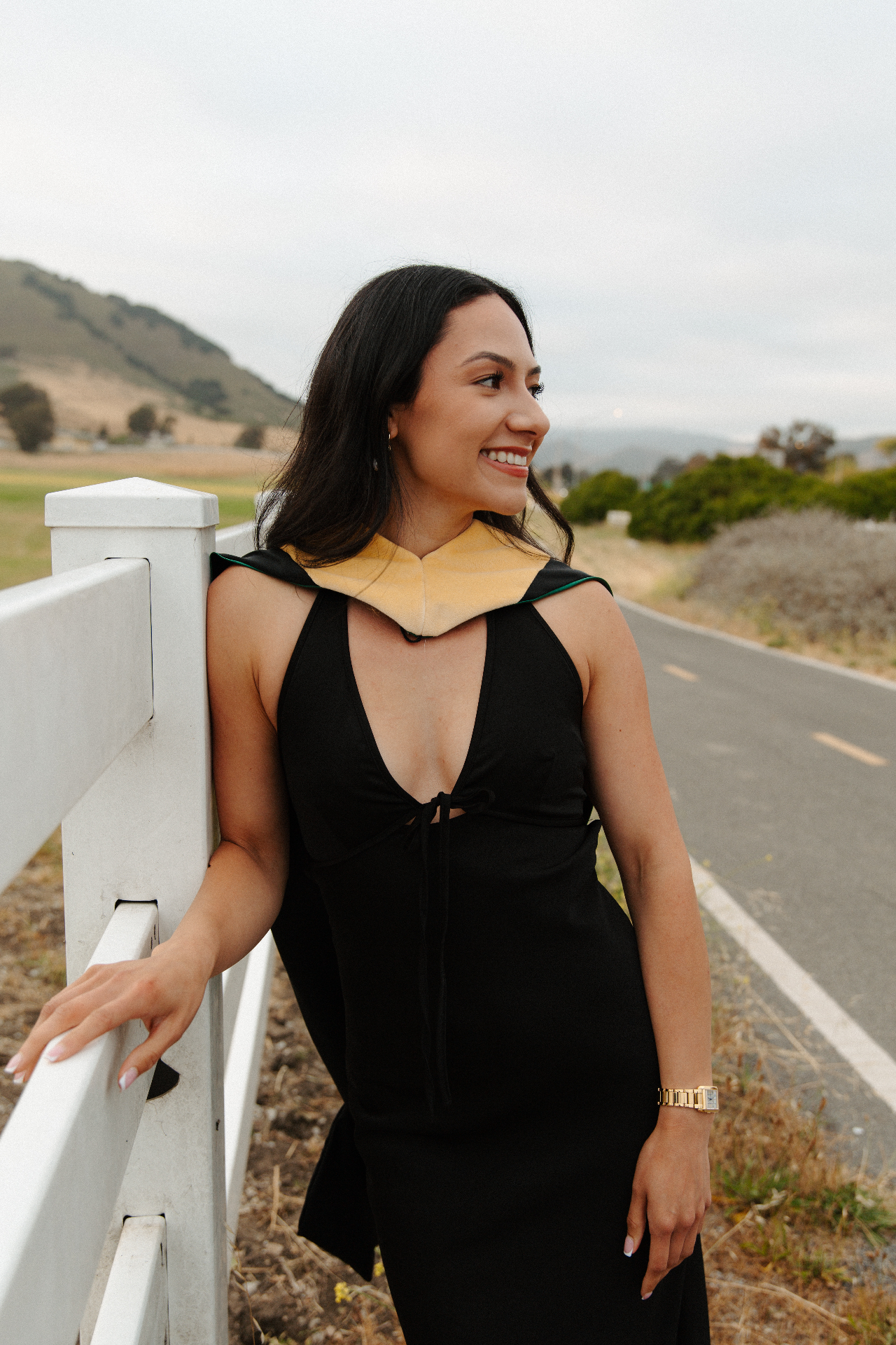 Graduate in a black dress and gold and green academic hood leaning on a white fence along a country road with rolling hills behind