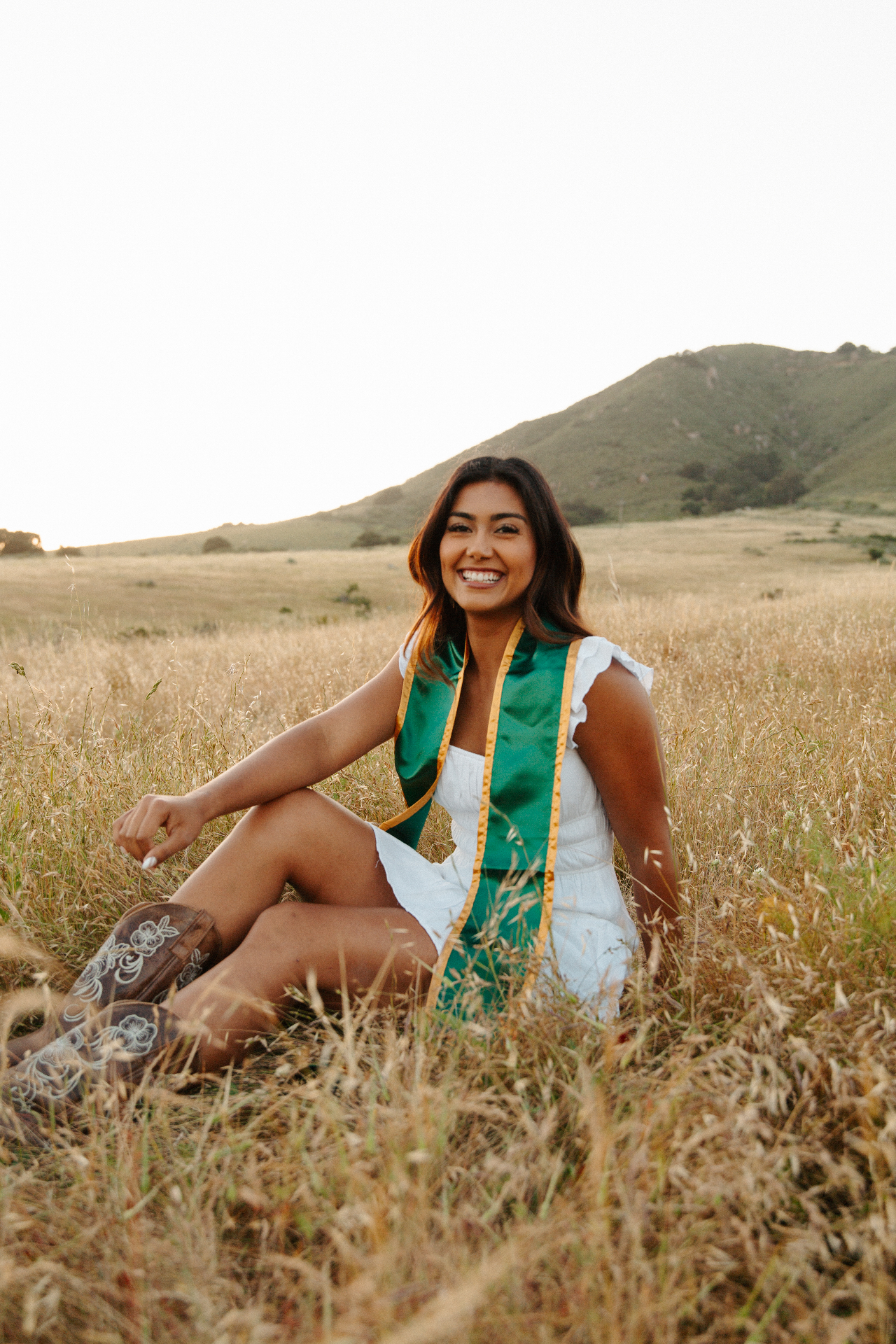 Graduate in a white sundress, cowboy boots and green and gold stole laughing while seated in a golden grassy field with rolling hills behind
