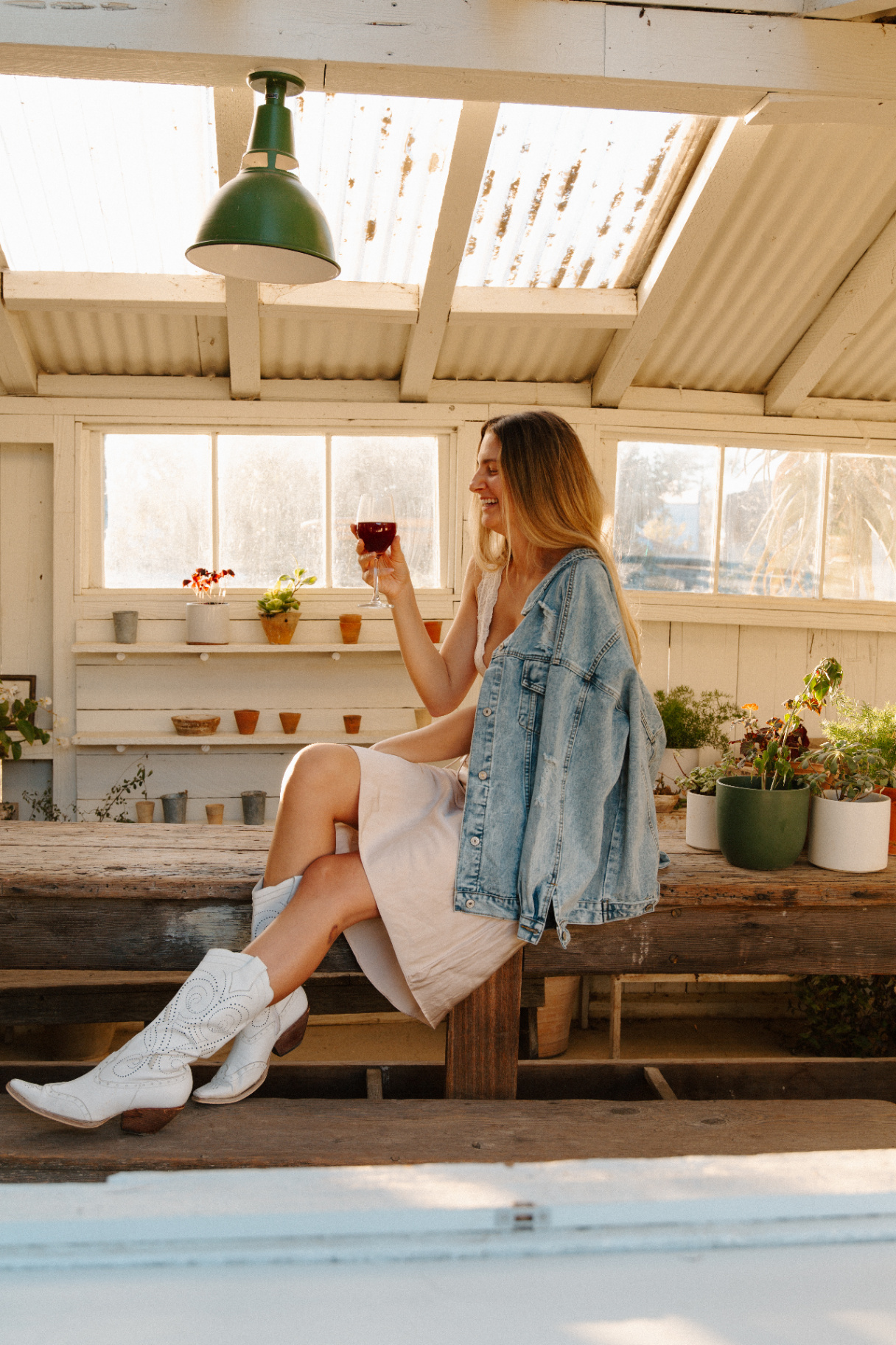 Woman in a slip dress, denim jacket and white cowboy boots laughing with a glass of wine on a wooden bench inside a sunlit greenhouse