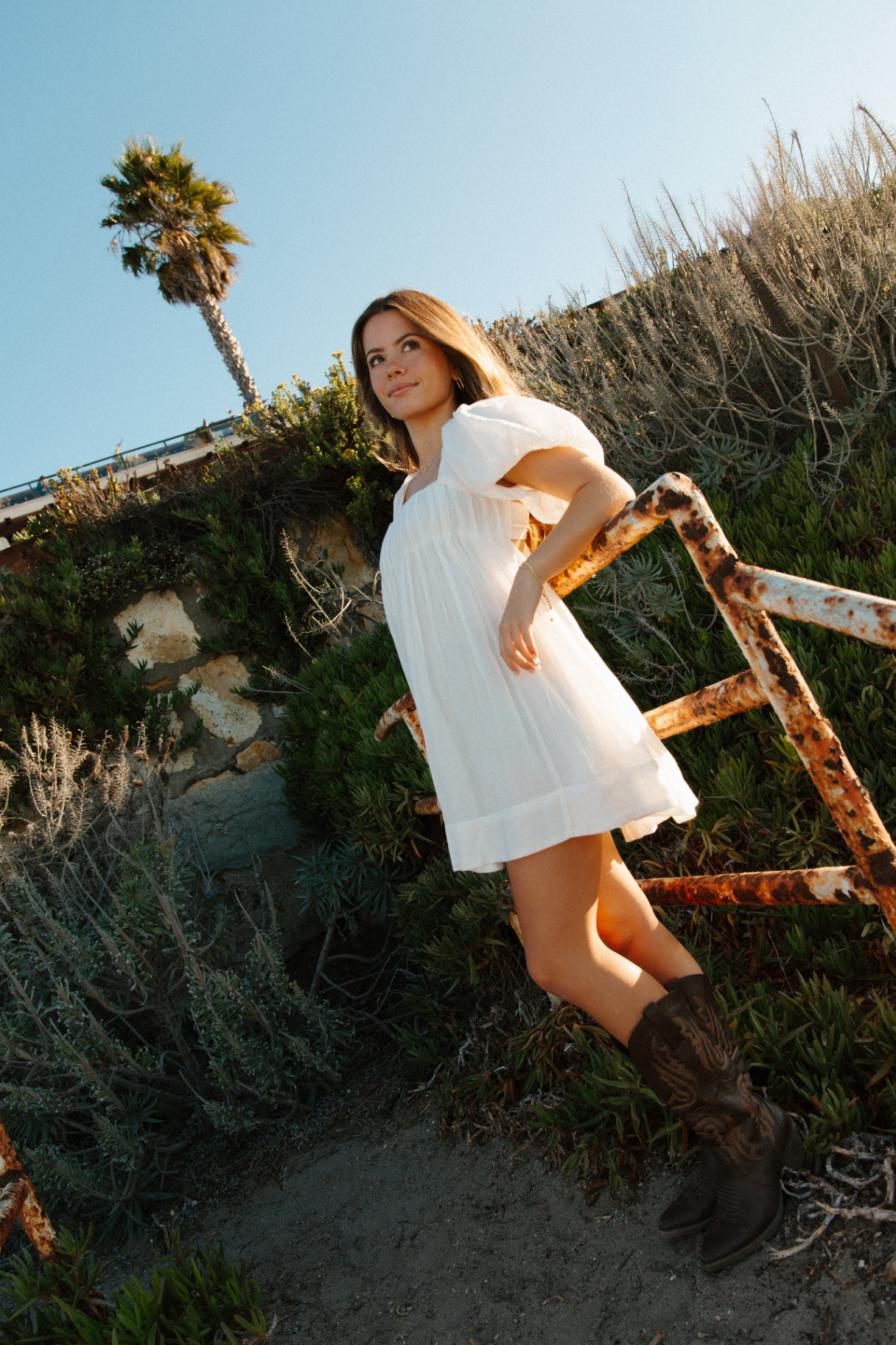Young woman in a white puff-sleeve dress and cowboy boots leaning on a rusted railing on a coastal hillside at golden hour