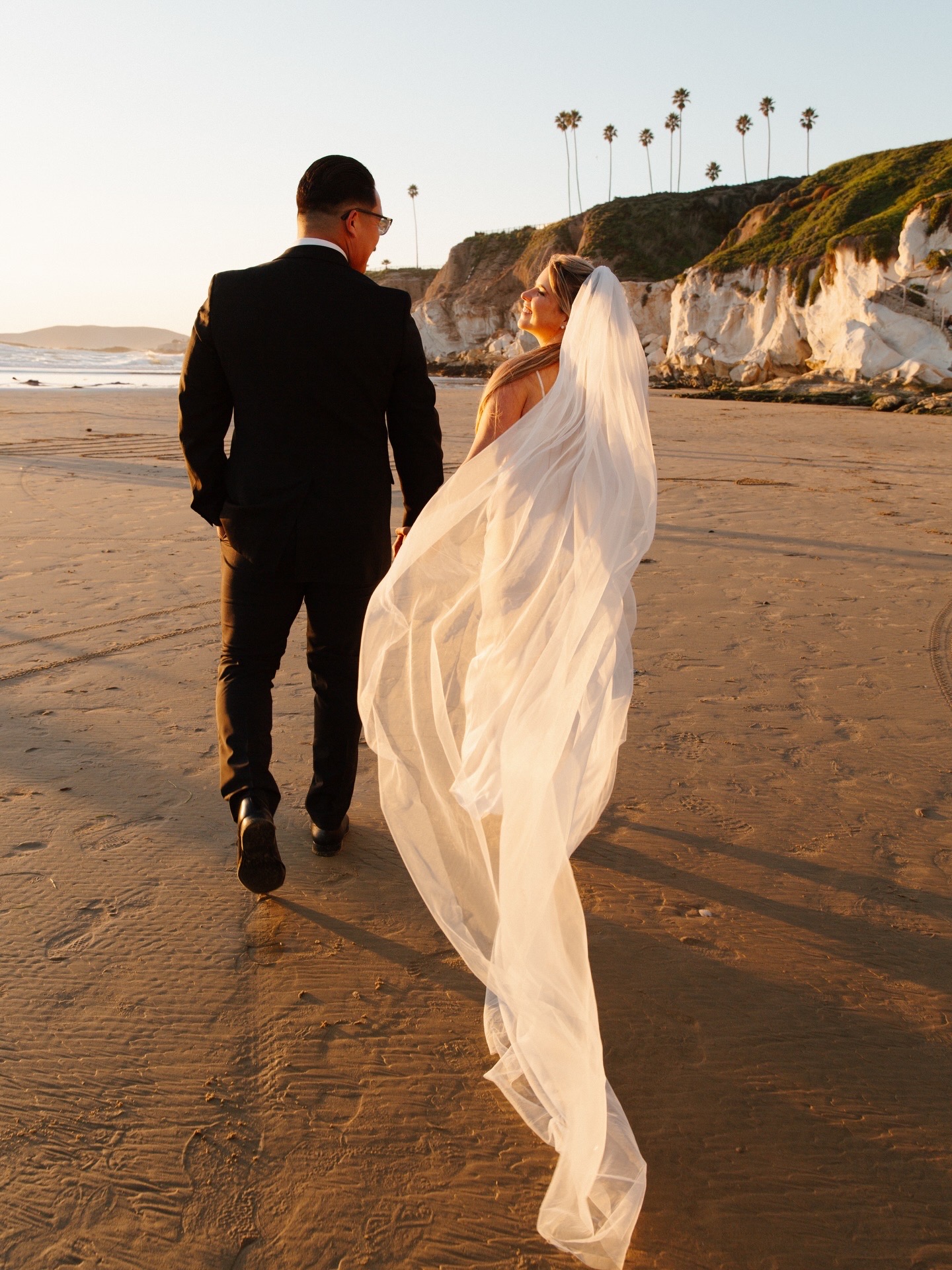 Bride and groom walking hand-in-hand on a sunset beach with a flowing veil