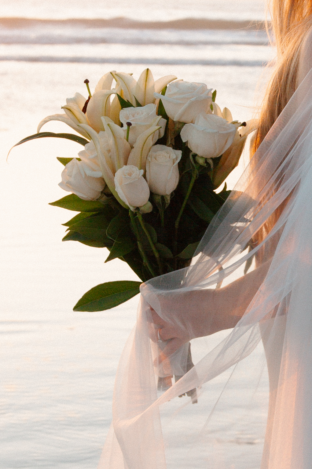 Bride at golden hour on the beach holding a white bouquet wrapped in veil