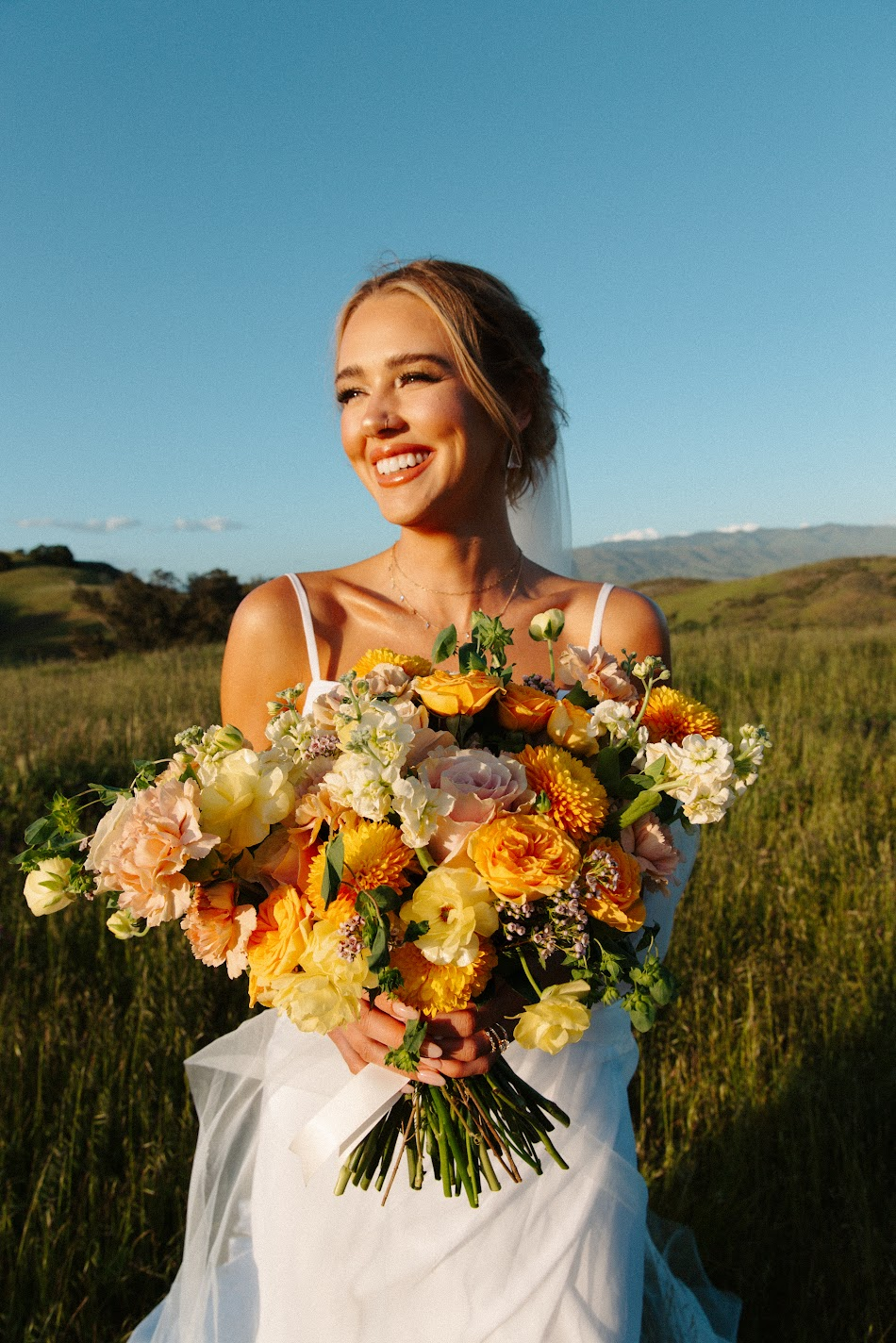 Smiling bride in a golden meadow holding a vibrant yellow and peach bouquet