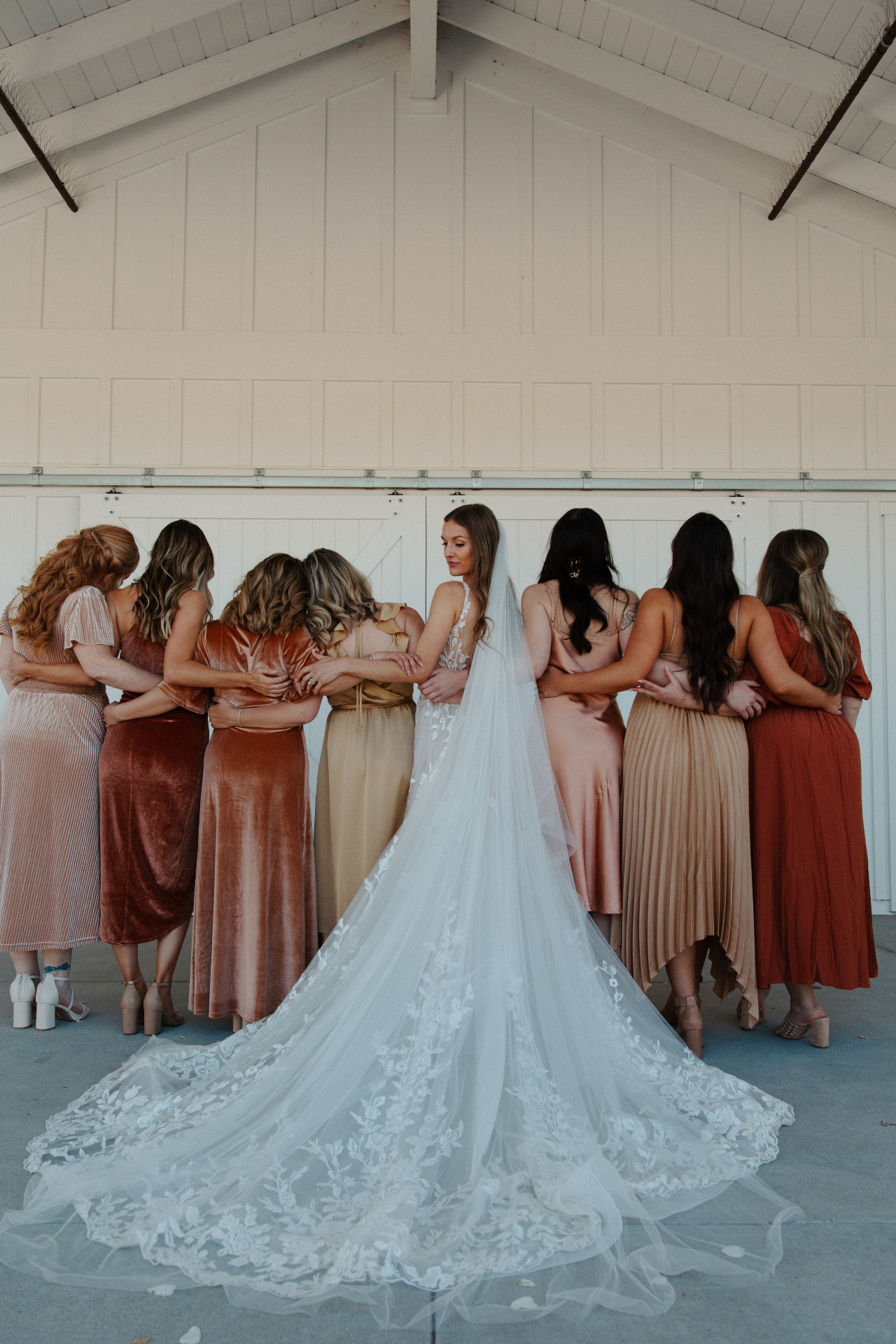 Bride with a long lace veil and train facing her bridesmaids in warm terracotta and champagne dresses