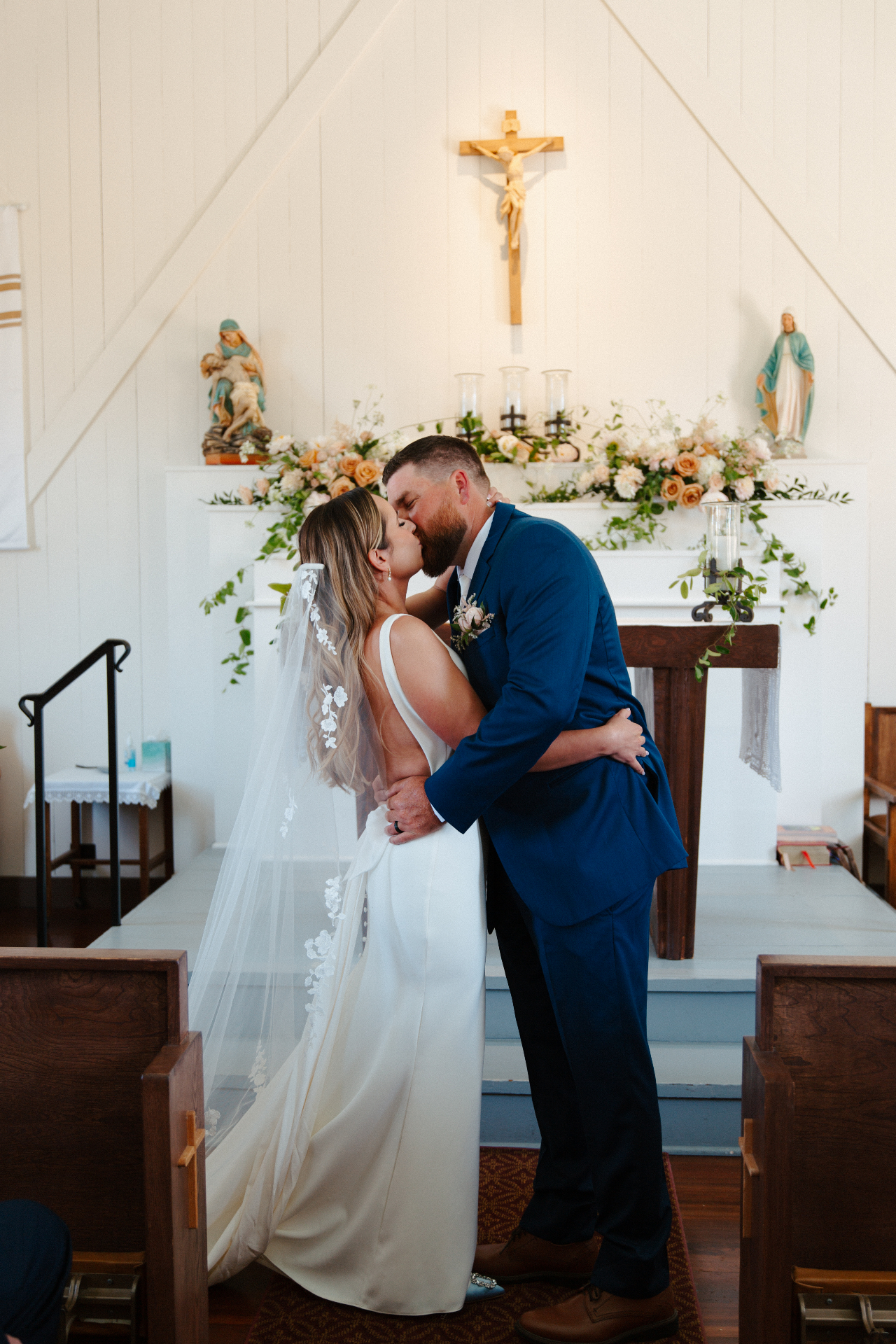 Bride in a long lace veil and groom in a navy suit sharing a kiss at a floral-draped chapel altar