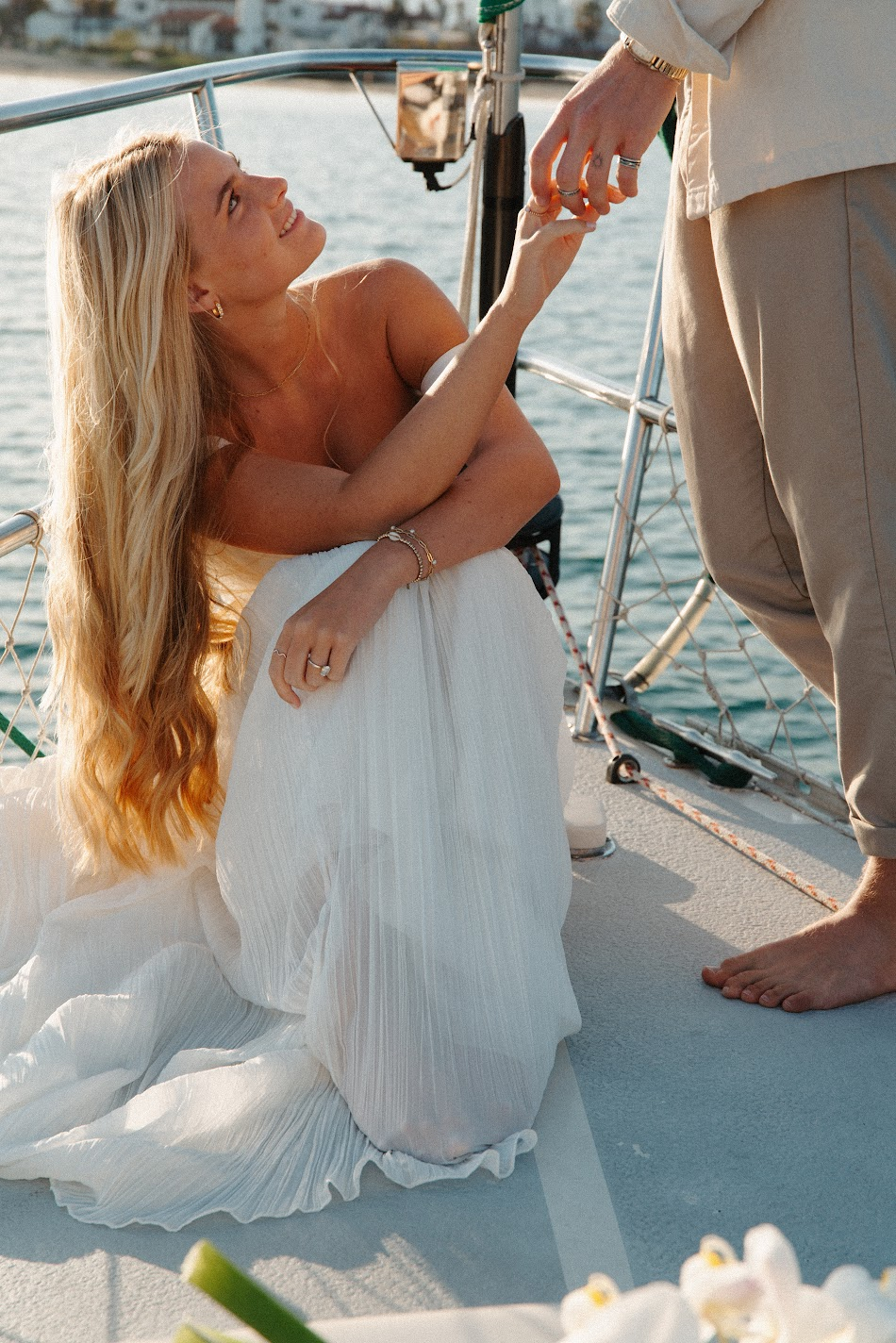 Bride seated on a sailboat at golden hour reaching up to her groom's hand