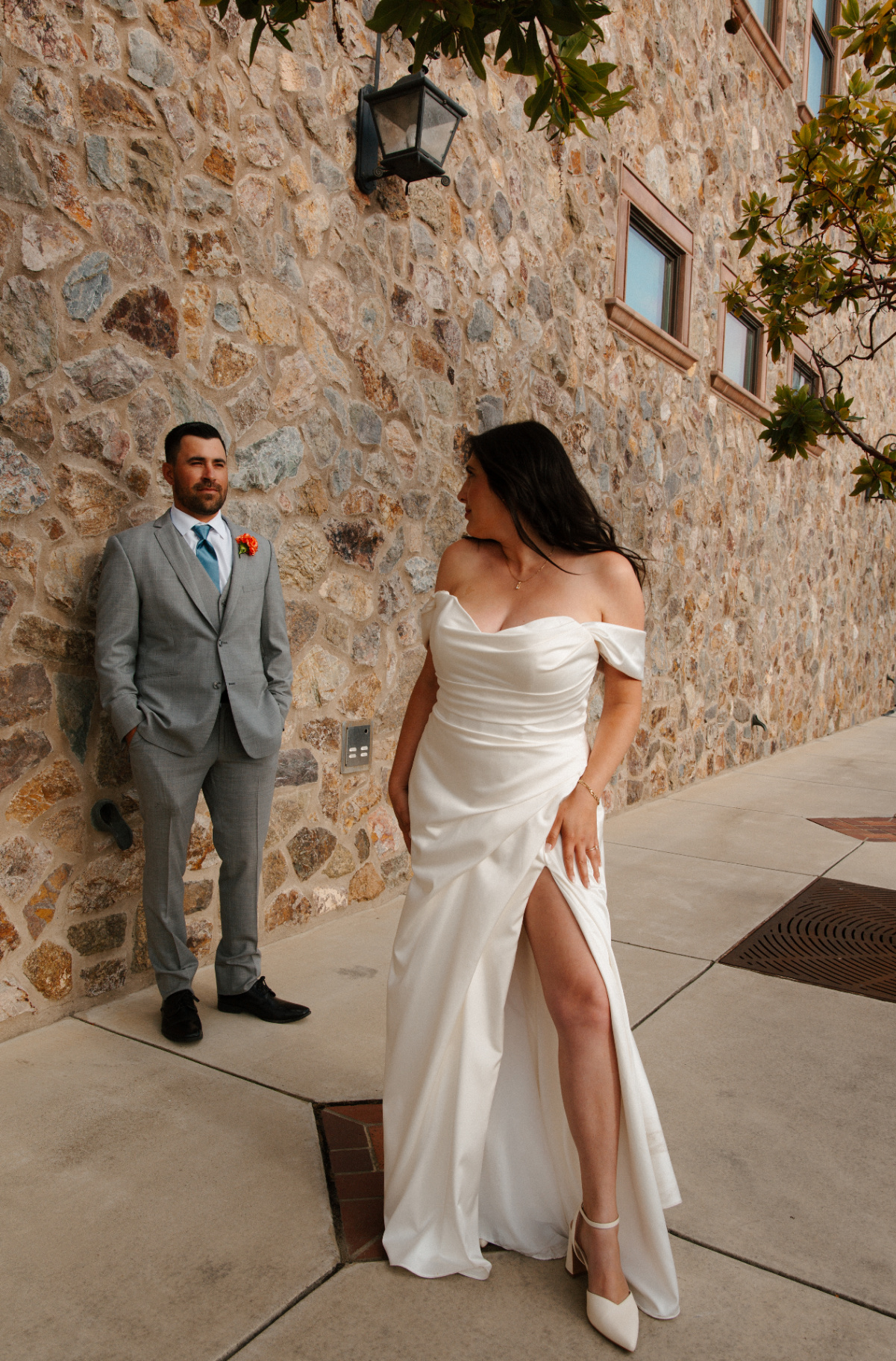 Bride in an off-the-shoulder satin gown with high slit glancing back at her groom in a gray suit and teal tie leaning against a stone wall