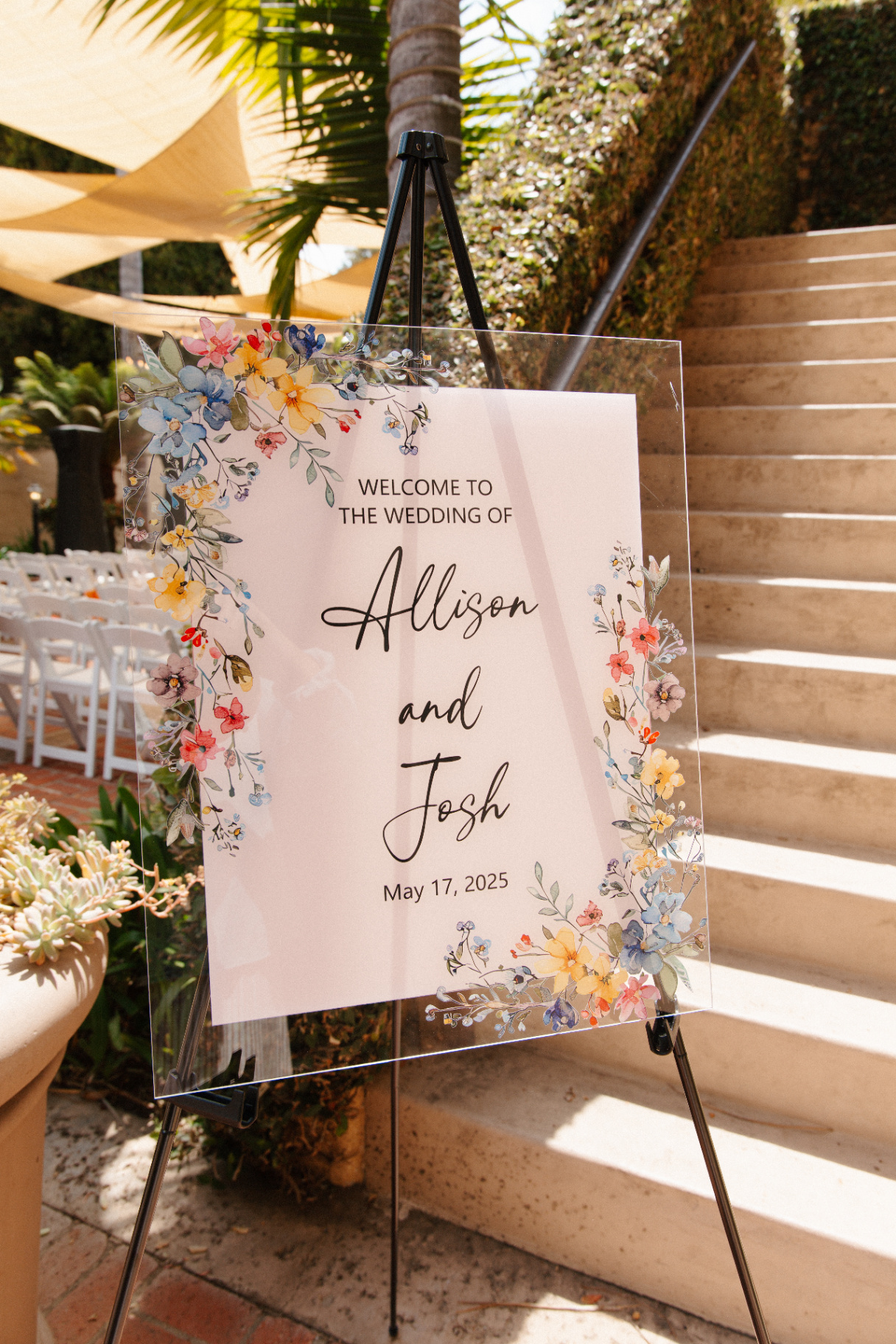 Acrylic wedding welcome sign for Allison and Josh with watercolor wildflower border on a black easel beside a garden ceremony aisle and stone steps